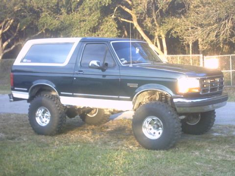 1992 Ford Bronco - Three-quarter front view of a dark green Ford Bronco with a white removable hardtop, lifted on a suspension kit with tall mud-terrain tires…