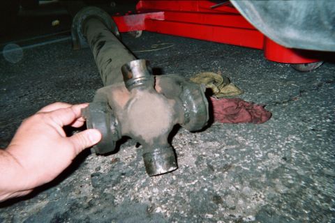 1990 Ford Bronco - hand holds a removed driveshaft showing its universal-joint cross on the pavement beside a red floor jack at night.