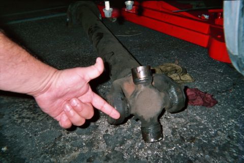 1990 Ford Bronco - finger points to the universal joint on a driveshaft lying on the pavement next to a red floor jack at night.