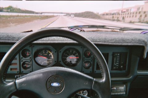 1985 Ford Mustang - Interior view of a car driving on a highway, showing the dashboard gauges and steering wheel.