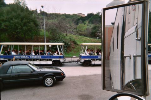 1985 Ford Mustang - A black convertible is parked near two Universal Studios tram tour vehicles carrying passengers.
