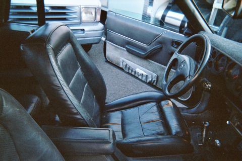 1985 Ford Mustang - Interior shot through the open driver's door of a 1980s Fox-body Mustang showing the black leather bucket seat with torn and worn bolsters,…
