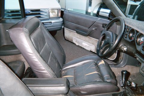 1985 Ford Mustang - Interior of a Fox-body Ford Mustang shot through the open passenger door, showing gray cloth-and-vinyl bucket seats with the passenger back…