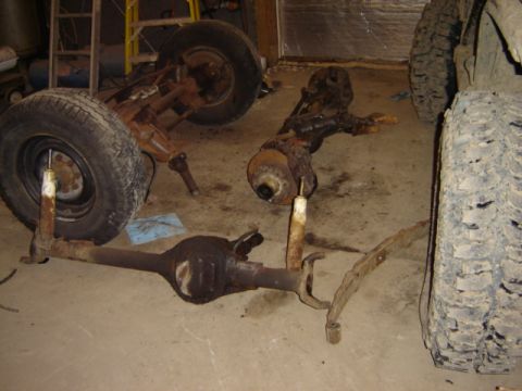 1992 Ford Bronco - Various vehicle axles and suspension components are laid out on a garage floor, with one axle supported by stands and a tire.