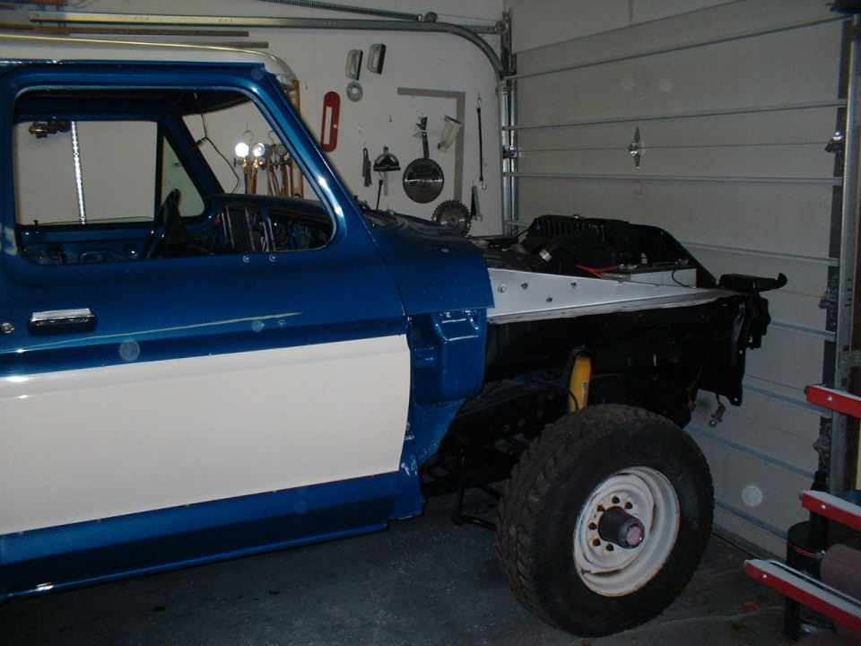 Pickup truck with its front end disassembled, parked in a garage. The cab is blue and white