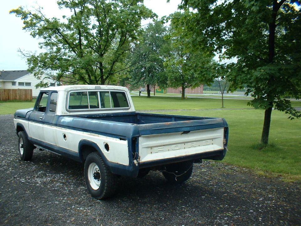 1978 Ford F-250 pickup truck, two-tone white and blue, parked on gravel with trees and a building in the background