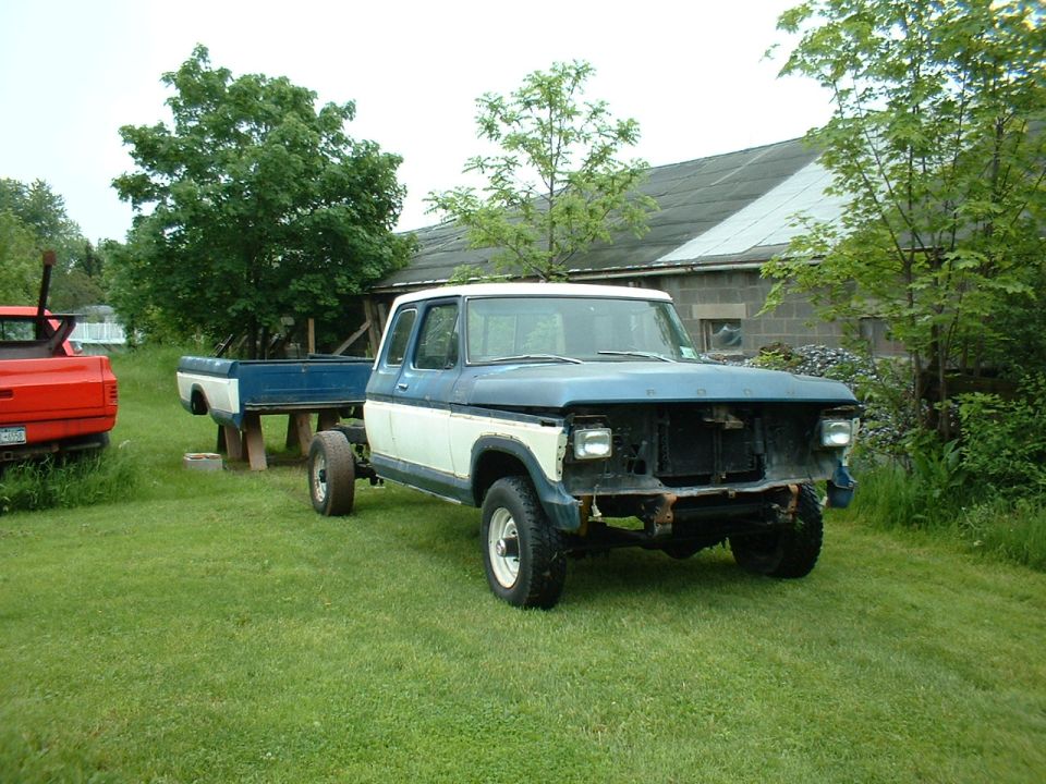 Ford F-Series truck cab and chassis, missing grille and bumper, parked on grass next to a red truck