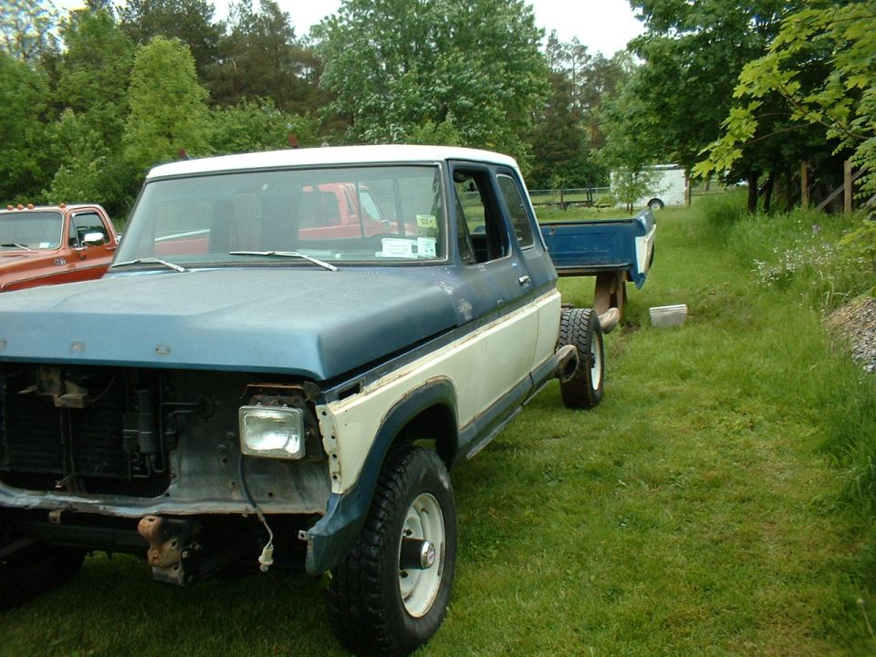 Blue and white Ford F-Series truck, missing grille and headlights, in a grassy yard