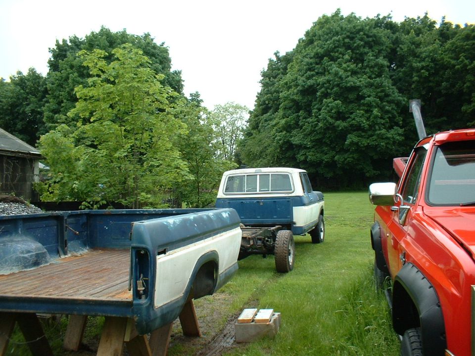 Blue and white pickup truck chassis with a red pickup truck next to it. A disassembled truck bed is in the foreground