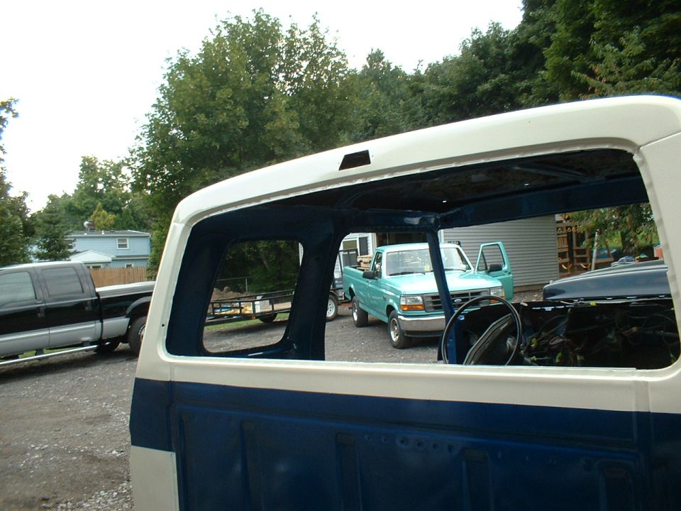 View through an open pickup truck cab shows a teal pickup truck with doors open and a black pickup truck in the background