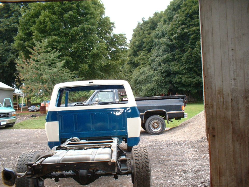 Truck chassis and blue-white cab, rear view. Another dark truck is parked behind it
