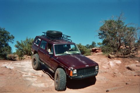 1991 Ford Bronco - Maroon Jeep Cherokee XJ with roof rack and spare tire on top, parked on rocky terrain under a clear blue sky.