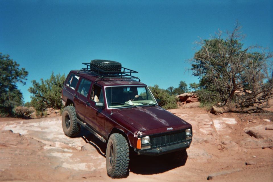 Maroon Jeep Cherokee XJ with roof rack and spare tire on top, parked on rocky terrain