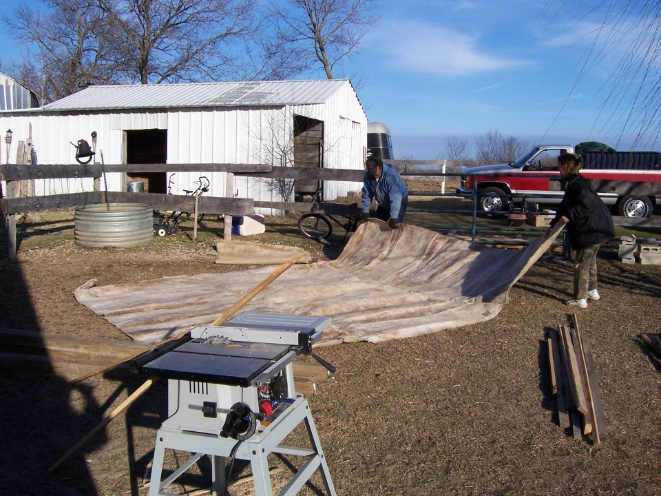 Two people are unrolling a large tarp in a yard with a red pickup truck parked in the background