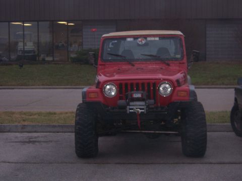 1999 Jeep TJ - This red Jeep Wrangler features a winch and oversized tires, parked in front of a commercial building.