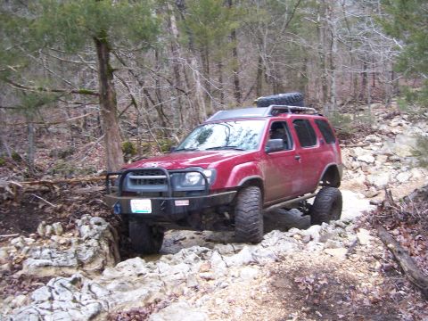 1999 Jeep TJ - This red SUV is navigating a rocky, wooded trail, demonstrating its off-road capabilities.