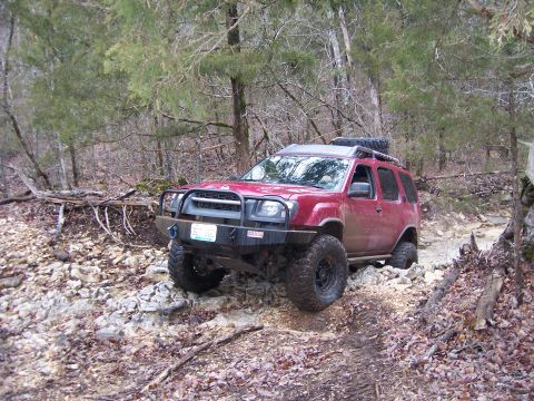 1999 Jeep TJ - A red SUV navigates a challenging rocky path through a wooded area.