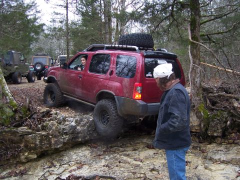 1999 Jeep TJ - A red SUV tackles an off-road obstacle, with other vehicles visible in the background.