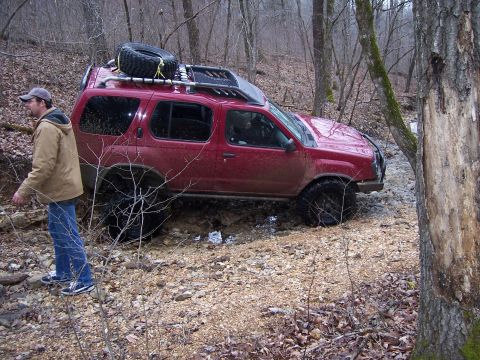 1999 Jeep TJ - A red SUV navigates a challenging off-road course in a wooded area, kicking up mud.