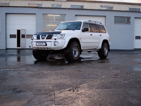 1996 Ford Bronco - A white Nissan Patrol SUV is parked on a wet asphalt surface in front of a grey building with garage doors.