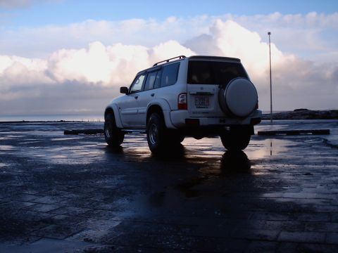 1996 Ford Bronco - A white SUV is parked on a wet surface near the ocean under a cloudy sky.
