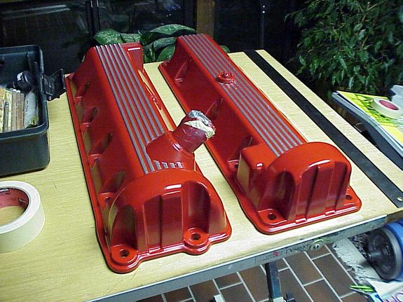 Two red powder-coated finned valve covers on a workbench, one tube masked with foil