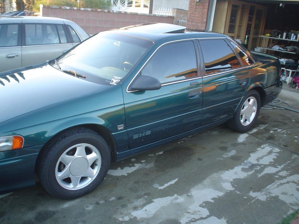 Wet dark green Ford Taurus SHO sedan, driver-side view on driveway by an open garage