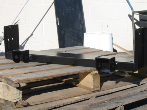 1992 Ford Bronco - A freshly painted black steel crossmember or bumper/frame mount bracket assembly with welded mounting tabs and bolt holes, resting on a woo…