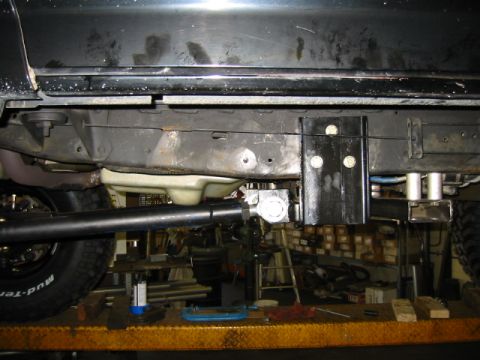 1992 Ford Bronco - Underbody view of a vehicle chassis showing a black fabricated steel mounting bracket bolted to the frame crossmember, with a driveshaft, d…