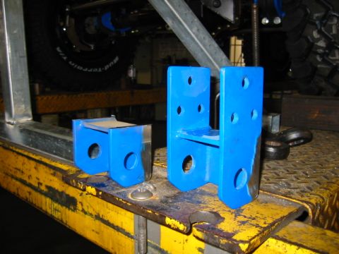 1992 Ford Bronco - Two freshly powder-coated blue steel brackets with multiple drilled bolt holes sit on a scuffed yellow work surface, apparently custom-fabr…