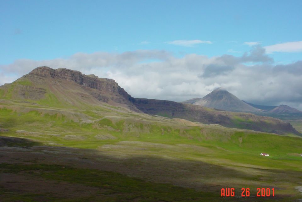 Vast green hills and mountains under a cloudy blue sky. Two small white buildings are visible in the distance on the right