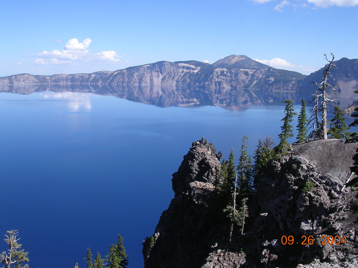 Deep blue crater lake ringed by cliffs and mountains with evergreens on a rocky point