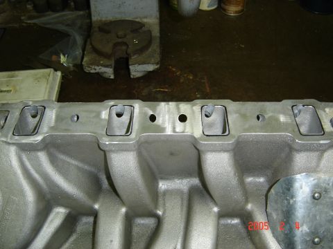 1990 Ford Bronco - Bare cast-aluminum V8 intake manifold sitting on a shop workbench, showing the machined port flange with rectangular intake ports, round co…