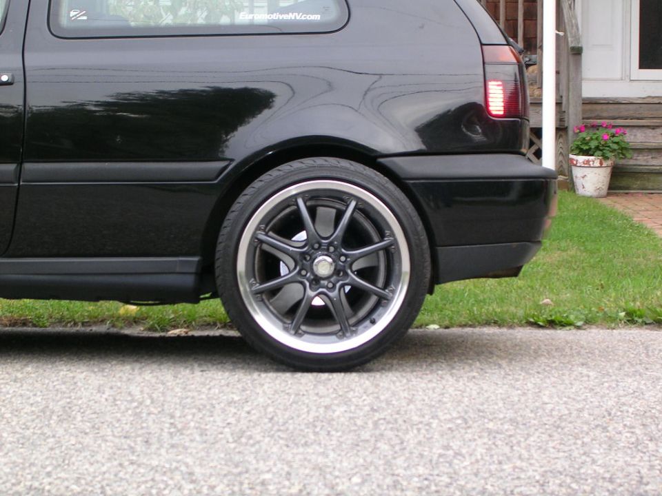 Black hatchback car rear quarter panel with aftermarket alloy wheel, parked on asphalt next to grass