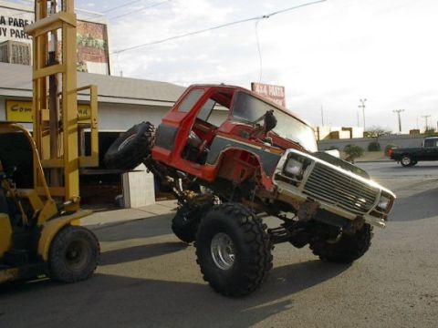 1985 Ford Bronco - A forklift lifts a heavily modified red truck, its grille indicating a 1980s model, while another truck is visible in the background.