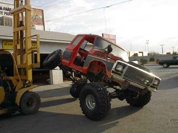 Red and gray lifted Ford truck on big mud tires, front raised by a yellow forklift at a 4x4 shop.