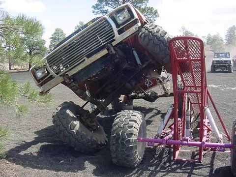 1985 Ford Bronco - An off-road vehicle is dramatically tilted, revealing its undercarriage and front grille, with another vehicle in the background.