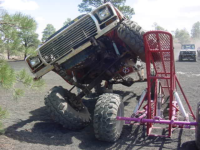Off-road vehicle tilted on its side, front grille and headlights visible, with a red metal structure in the foreground