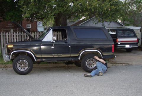 1988 Ford Bronco - Person works on a black 1988 Ford Bronco while another Bronco is parked nearby.