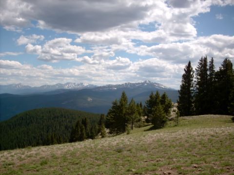 1986 Ford Bronco - A vast mountain vista unfolds under a dramatic sky, with dense pine forests covering the slopes.