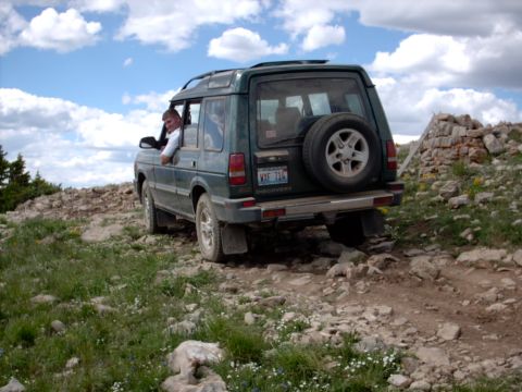 1986 Ford Bronco - A green SUV navigates a challenging rocky terrain on a mountain road under a cloudy sky.