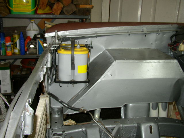 Engine bay showing a yellow-top Optima battery secured with a strap, with metal sheeting and various hoses visible