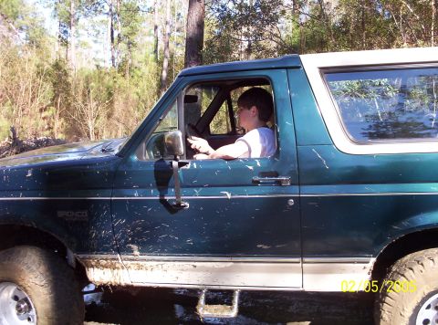 1993 Ford Bronco - A muddy 1993 Ford Bronco is driven by a person through a wooded area.