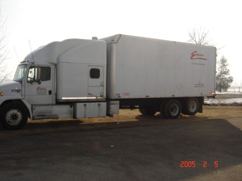 1990 Ford Bronco - A white semi-truck and trailer are parked on a paved surface with some dry grass and trees in the background.