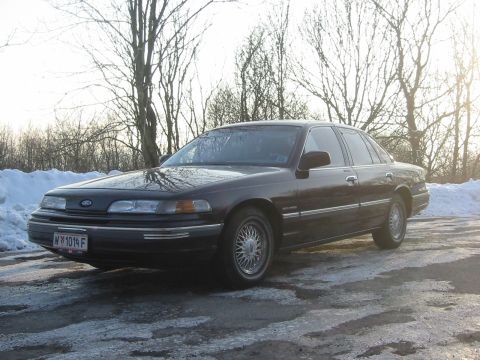 1992 Ford Crown Victoria - A dark sedan is parked on a road with snow and ice on the ground, with trees in the background.