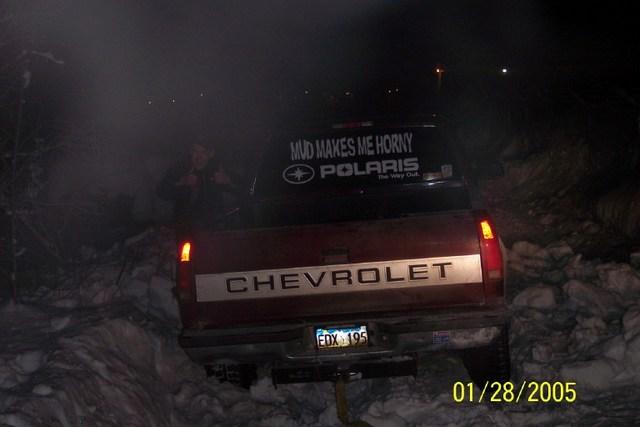Rear view of a red Chevrolet pickup truck with a sticker that says 'MUD MAKES ME HORNY POLARIS'. The truck is in a snowy area