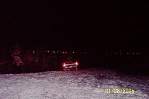 1981 Chevrolet C/K Pickup - A pickup truck is seen from the rear, driving on a snow-covered area at night with distant lights in the background.