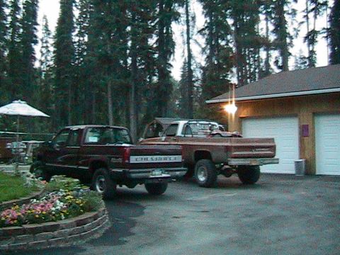 1981 Chevrolet C/K Pickup - Two Chevrolet pickup trucks are parked in front of a garage with trees behind them.