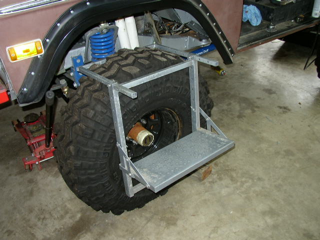 Close-up of a large off-road tire with a custom metal step attached to its side. A red hydraulic jack is visible underneath the vehicle's fender