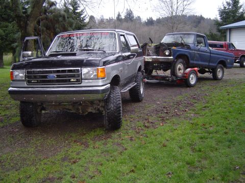 1979 Ford Bronco - A lifted black and silver SUV tows a blue pickup truck on a trailer, with "4x4" visible on the SUV's fender.